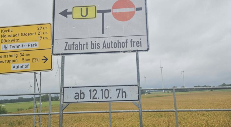 Hinweisschild an der B 167 mit Pfeil nach links und Umleitungstafel „U“, Verbotsschild für die Gegenrichtung, Text „Neuruppin – Zufahrt bis Autohof frei“ und Zusatzschild „ab 12.10. 7h“; links daneben ein gelber Wegweiser mit Kyritz, Neustadt (Dosse), Bückwitz, Temnitz-Park und Neuruppin 5 km, im Hintergrund Windräder