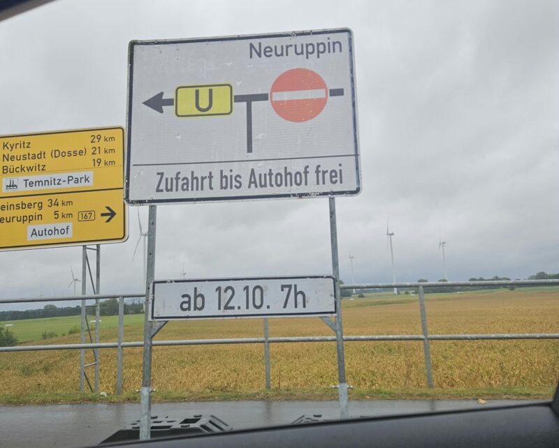 Hinweisschild an der B 167 mit Pfeil nach links und Umleitungstafel „U“, Verbotsschild für die Gegenrichtung, Text „Neuruppin – Zufahrt bis Autohof frei“ und Zusatzschild „ab 12.10. 7h“; links daneben ein gelber Wegweiser mit Kyritz, Neustadt (Dosse), Bückwitz, Temnitz-Park und Neuruppin 5 km, im Hintergrund Windräder